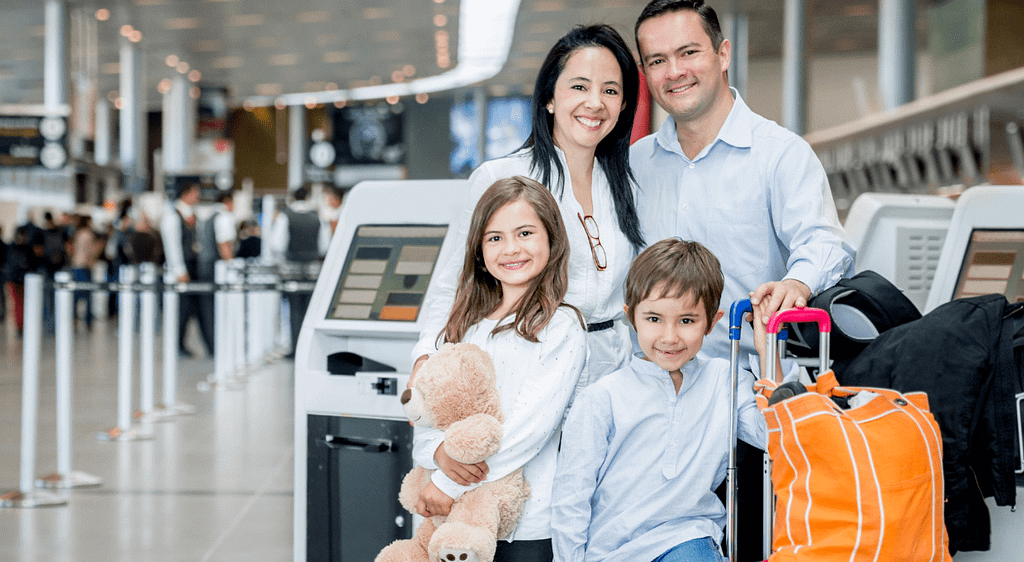 Smiling family with two children holding a teddy bear, standing in an airport terminal with luggage ready for their trip.